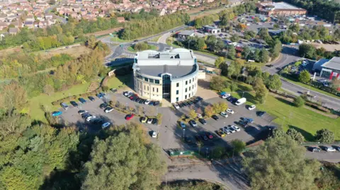 An aerial, drone-style photograph of a modern circular building surrounded by parking areas, greenery, and a mix of urban and suburban elements