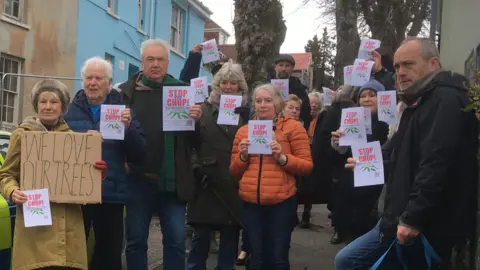 BBC Campaigners gather on the pavement next to the three trees that are due to be felled. There is more than ten people there along with two dogs. They are holding paper leaflets saying "Stop the Chop" and a cardboard placard reading "We love our trees." There is a police car parked in the road some council workers in the background. 