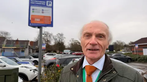 BBC News/Jasmine Lowe A man with white hair in a suit, standing in a car park next to a large sign where there is small print with instructions on how to park 