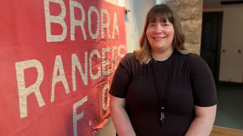 Aileen Ogilvie is standing inside Brora Heritage Centre next to a red cloth banner with the words "Brora Rangers FC" stitched on it in white letters. She has shoulder length brown hair and is wearing a black top. She is smiling.