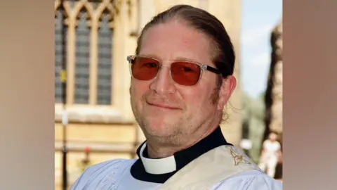 A man with long hair that has been tied back. He is wearing glasses that are tinted red and he wears clergy clothing.