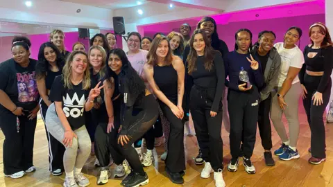 BBC A group of around 20 women are standing together and smiling. They are wearing leggings and tops with some holding water bottles. Bright pink lights shine in the background of a dance studio