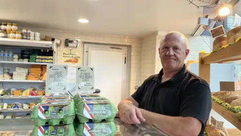 A market trader standing by a lot of cheese, eggs and bread