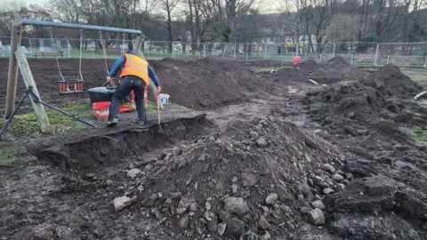 A man digs in front of a pair of swings. Around him are large mountains of earth in a play park which has been almost completely dug up 