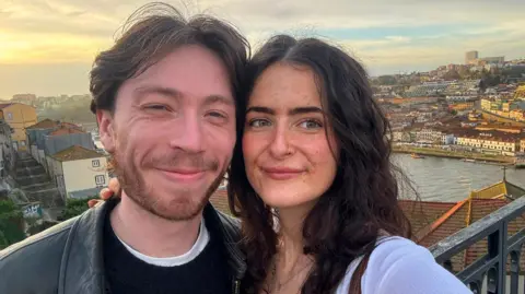 Gracie A smiling young couple with dark hair take a selfie at a scenic viewpoint, with buildings and water visible in the background.