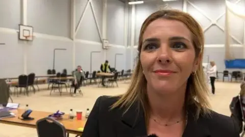 A woman with brown/blonde hair is on the right of the photo wearing a dark jacket. She is standing in a sports hall.