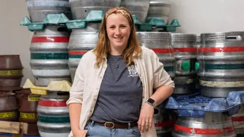 Kate Balchin A woman stands in front of a stack of beer kegs, leaning on them. She has pale skin, light brown hair, and is smiling. She's wearing an orange and brown hairband, a grey t shirt with 'Wold Top' written on it, a light coloured button up shirt over the top, blue jeans, and a brown belt. She is also wearing a smart watch.