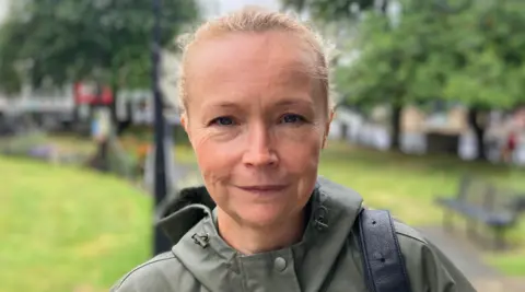 Helen is looking directly at the camera and smiling in this tightly-cropped image. She has blonde hair tied back and is wearing a green mac/waterproof with a bag over her shoulder.