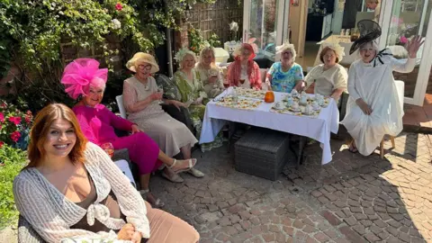 Paula Gamester with the other ladies, all dress in their finery, sitting round a table in the garden smiling at the camera