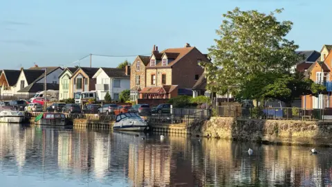 Dr Syntax A range of houses, some semi-detached, with a large tree to the right, reflected in a river, shot from the opposite bank, with the water disappearing into the foreground.