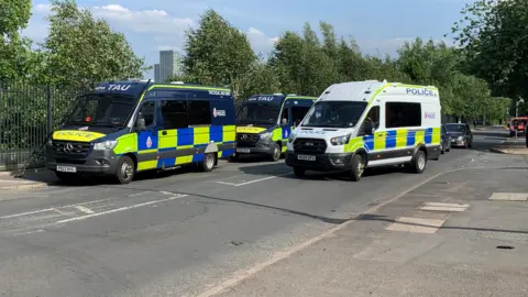 Phil Cunliffe/BBC Three police vans are parked blocking a road. Cars are driving behind them.