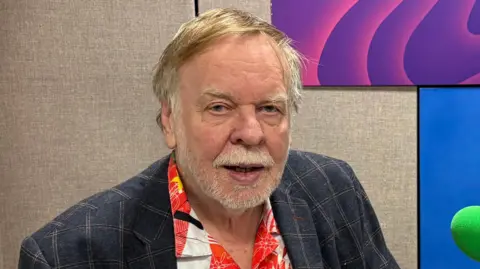Johnnie Wright/BBC A man smiles at the camera as he sits in a radio studio. He has short hair – some blond and some is grey. He is wearing a navy suit jacket with a Christmas-themed shirt underneath. There is a green microphone on the right.
