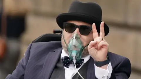 PA Media A man in a pin-striped suit and bow tie with trilby hat and sunglasses gives a victory salute