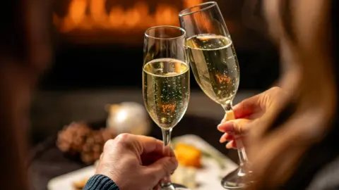 Getty Images Um casal mais velho brindando duas flautas de champanhe em sua sala de estar