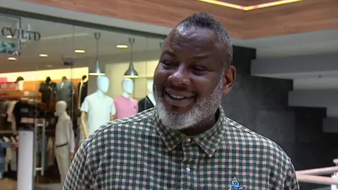 Mark Barratt - a man with short balck hair and a grey beard is wearing a chequered shirt and smiling. In the background, a clothing shop is visible