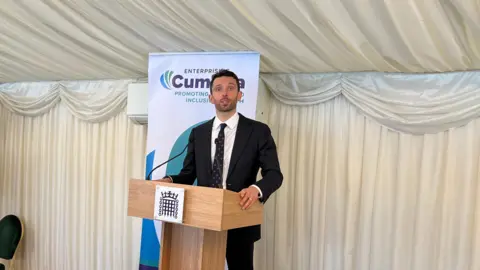 MP Josh MacAlister speaking at Cumbria Day from a lectern. He has short black hair and is wearing a black suit jacket and trousers with a white shirt and navy blue tie with range circles.