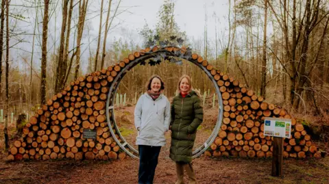 Artist Lucy Pittaway, wearing a green coat, stands next to estate owner, Felicity Cunliffe Lister, in a white coat, in front of an archway of logs at the entrance to the new woodland.