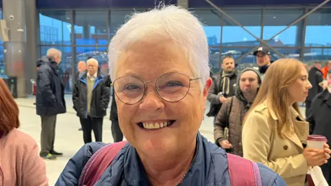 A woman smiles into the camera. She's wearing glasses and short grey hair. She's wearing a navy coat and has a red rucksack over her back. There are a lot of people standing behind her.