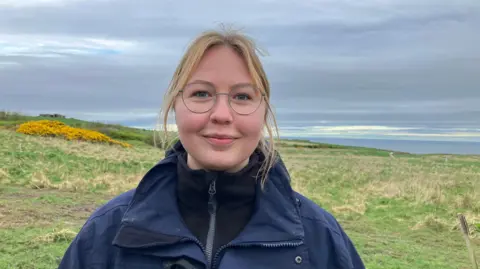 A young woman with blonde hair tied back, wearing glasses and a black polo neck fleece with a navy waterproof jacket on top. She is smiling and standing in a field slightly away from the cliff's edge.