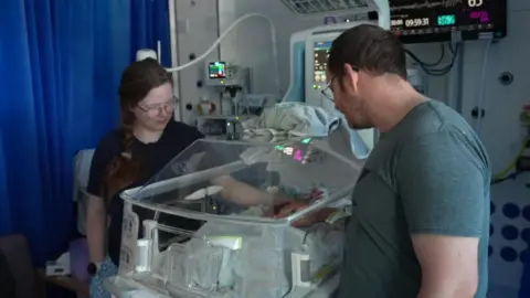 a Mother and father stand beside an incubator containing their new baby in a neonatal ward 