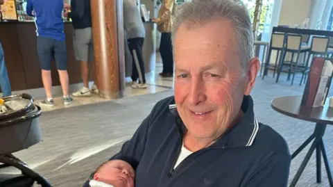James Messhamsat in a chair in a restaurant holding a baby