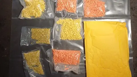 Clear plastic bags containing hundreds of yellow and orange pills are laid out on a plastic sheet. A brown envelope is lying next to the bags. 