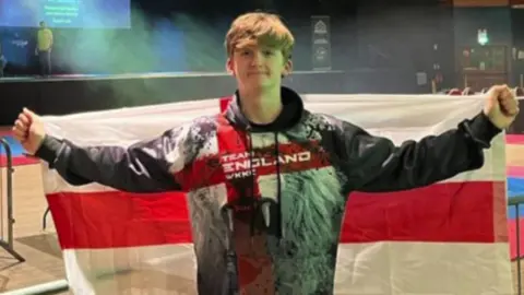 Alex Eastwood is holding an England flag behind his back. He is staring and smiling directly at the camera. He is wearing a hooded jumper with a lion and the words "Team England" written in white on top of a red cross. 
