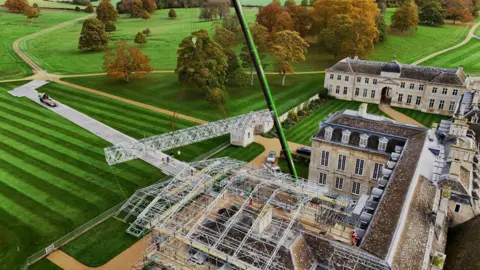 County Lifting A large stately home with extensive restoration work being carried out. There are lawns around the building with trees in the distance. There is scaffolding to the right and a large green crane. 