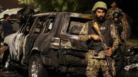 Reuters A security forces officer in helmet and military garb stands in front of a scorched vehicle holding a rifle