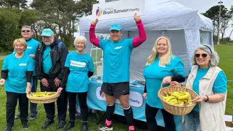 Ryan O'Shea A group of volunteers in turquoise shirts stand in front of a white gazebo with The Brain Tumour Charity branding, holding baskets of bananas, with a table of information materials set up on a grassy field.