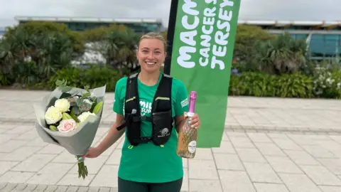 Daisy is standing outdoors holding a bouquet of flowers and a bottle of sparkling wine, wearing a green 'Macmillan Cancer Support' shirt and a black vest. Behind her is a large green flag that reads 'Macmillan Cancer Support Jersey,' with greenery and buildings in the background