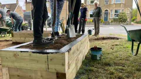 Tom Jackson/BBC On the left are raised flower beds which are made from wood. Children are standing on the soil and are holding spades. There are people in the background watching.