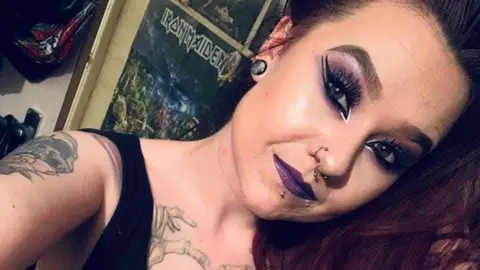 Simone Smith is taking a photo of herself. She is wearing eyeliner, purple eyeshadow and matching purple lipstick. She has tattoos, her nose, lip and ears pierced, and there is an Iron Maiden poster in the background.  