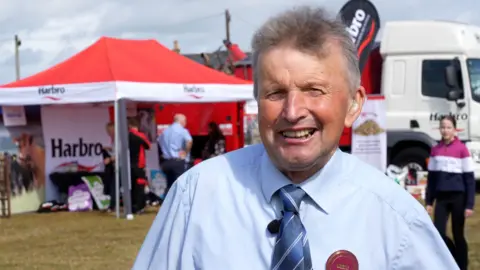 A man with spiky greyish/black hair in a light blue shirt with a blue striped tie stands in front of an agricultural show field
