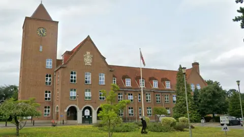 A red brick office building. the building has rows of windows and a crest over the front entrance. A clock is in the centre of a tower on the left of the building. There is a garden with trees, flagpoles and a black metal figure of a soldier in front of the building.
