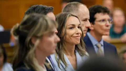 Karen Read smiles as the not guilty verdict is read during her trial. She is wearing a light blue suit and is flanked by her legal team.