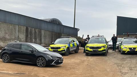 Cars and police vehicles sit on a dirt path near farm buildings