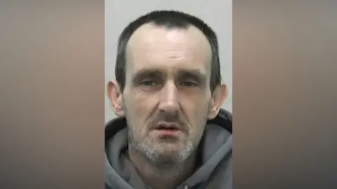 Mugshot of Young. He is a dishevelled looking man with pale pasty skin, a glazed expression on his eyes. He has grey stubble and short black hair.