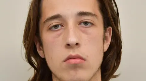 Alfie Coleman has shoulder-length brown hair swept backwards. He looks tired but shows no emotion while pictured in this custody mugshot.