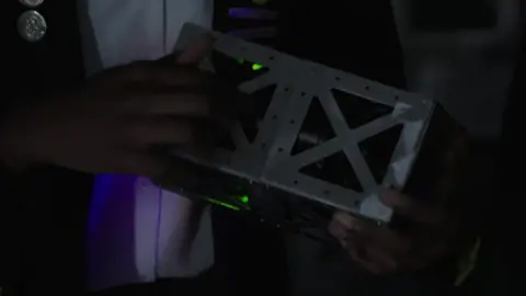 A close up of the satellite's metal casing - it is held in the hands of one of the pupils in a darkened room and it has some small green lights on the inside of the casing. 