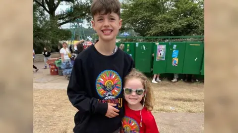 Family handout Kelly's children with their arms around each other in front of a bank of porta-loos. Her daughter is in a red Glastonbury top and sunglasses, and her son is in the same top in black.