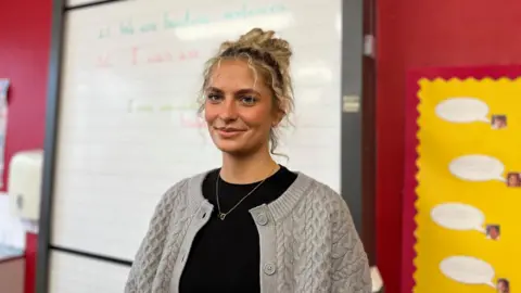 Isla wearing a black top and a light grey cable‑knit cardigan stands in a classroom. Behind them is a large whiteboard with handwritten text in green and red marker. To the right is a yellow classroom display board with speech‑bubble cutouts, and to the left is a red wall with part of a dispenser visible. The setting appears organised and brightly coloured.