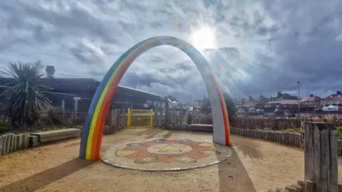 BBC Weather Watchers / MackemIan A children's playground, surrounded by a small log fence with a yellow gate. In the centre is another circle with coloured shapes and numbers, and an arched rainbow structure over it. There are houses and a large but low building behind it, and the sky is cloudy with sun producing an almost prism effect through a gap.