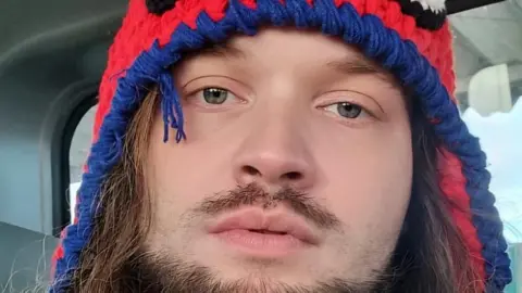 Wirley Contaifer A man with brown facial hair looks at the camera wearing a Spider-Man hat 