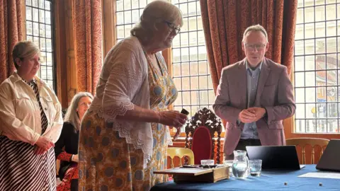 Royal Surrey County Hospital Anne Hope, Jenny Watkins and a man in a suit jacket observe two small dice on a table in an ornate, wood-panelled room. A woman in mayoral chains is sat in a chair behind them.