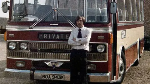 Grettons Coaches Roger Gretton standing in front of a coach, wearing a shirt and black tie