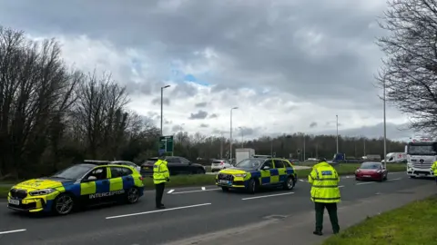 A road with two police cars parked in the middle. There are two police officers standing on the road, as well as a number of other vehicles. The sky is cloudy with patches of blue. 