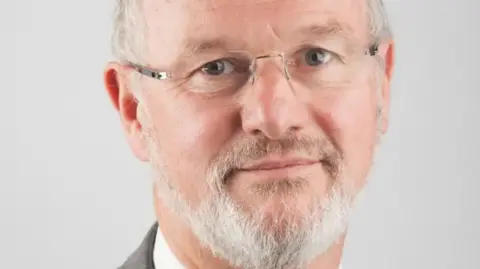 A headshot of Stephen Davies, who had a white beard and glasses, is looking at the camera in front of a grey background.
