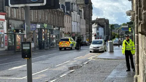 Armed police beside a police car in the middle of a street. There is a police officer in a hi-vis jacket in the foreground looking at the camera 