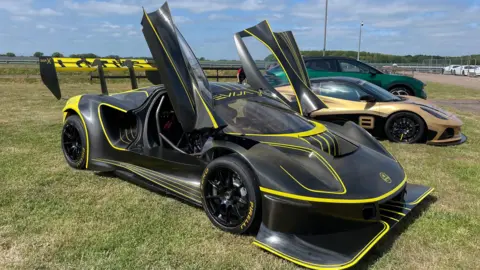 Edd Smith/BBC A black and yellow sports car is parked on grass with its doors open alongside other cars